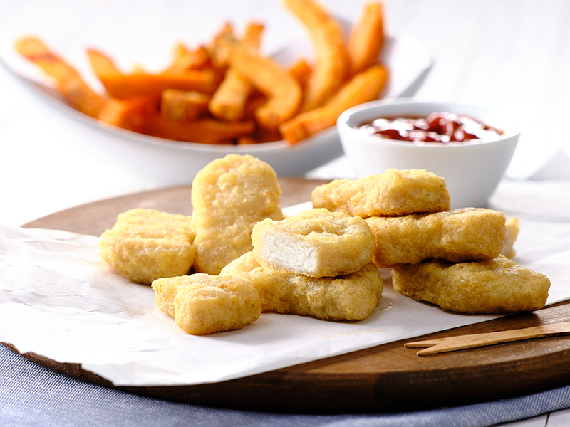 Chicken Nuggets auf einem Holzbrett mit Süßkartoffelpommes im Hintergrund.